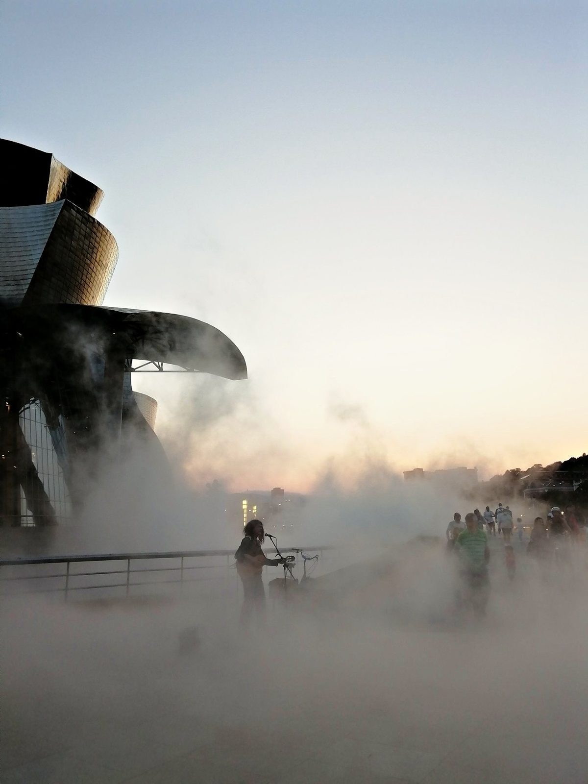 Un músico y paseantes entre la niebla junto a un edificio de titanio